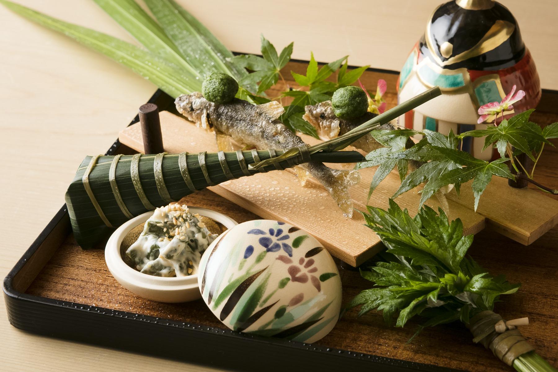 5月の献立から。清々しい香りの「菖蒲八寸」。兜の中にはトゲグリ蟹、白ダツ芋の黒酢ゼリー。尾形光琳写しの香合の中には、干しぜんまい白和えが入る。緑酢おろしを頭にのせた稚鮎唐揚げ、鯛の粽寿しが爽やかな風を運ぶ。